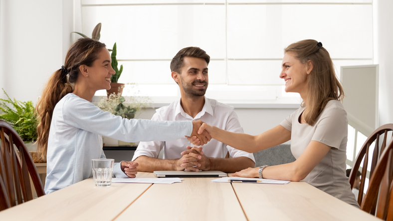 Happy female business partners shake hands sign two contracts with lawyer mediation at meeting, smiling businesswomen handshake make legal partnership deal after successful negotiations with mediator